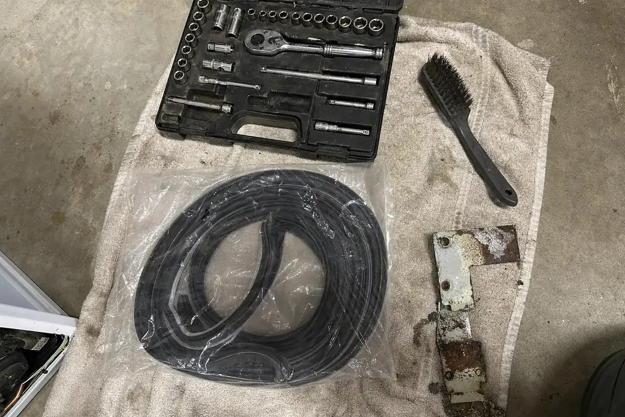 Overhead flat-lay on a weathered towel of a socket set, a wire brush, a bagged door gasket and a corroded bracket