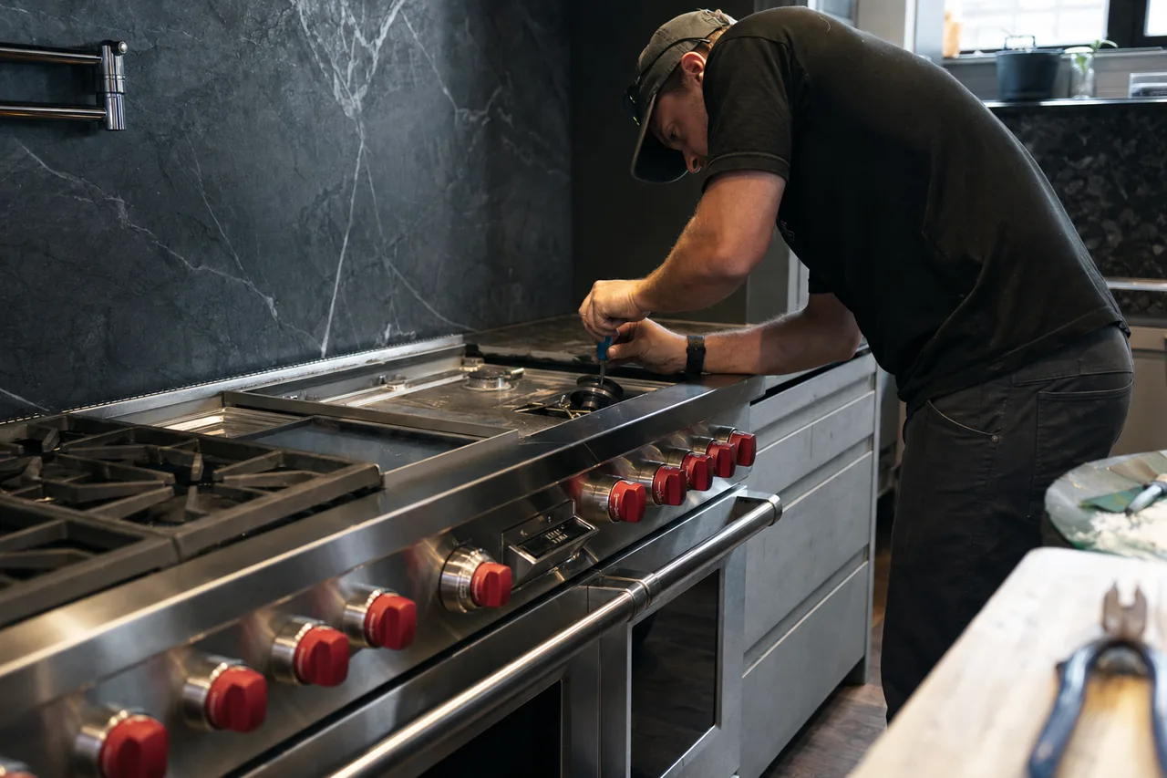 A stainless professional range in a bright kitchen with a grate removed and a technician working at the burner head